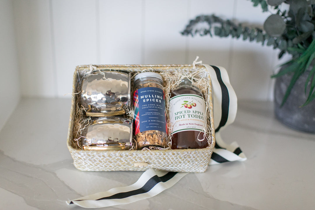 Gift basket with spiced apple hot toddy cocktail mix, mulling spices, and set of silver mule mugs set on a white surface