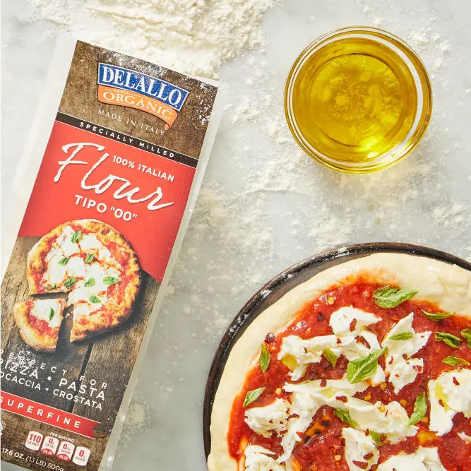 Arranged on a white countertop is a bag of flour as well as a cup of oil and a pizza.