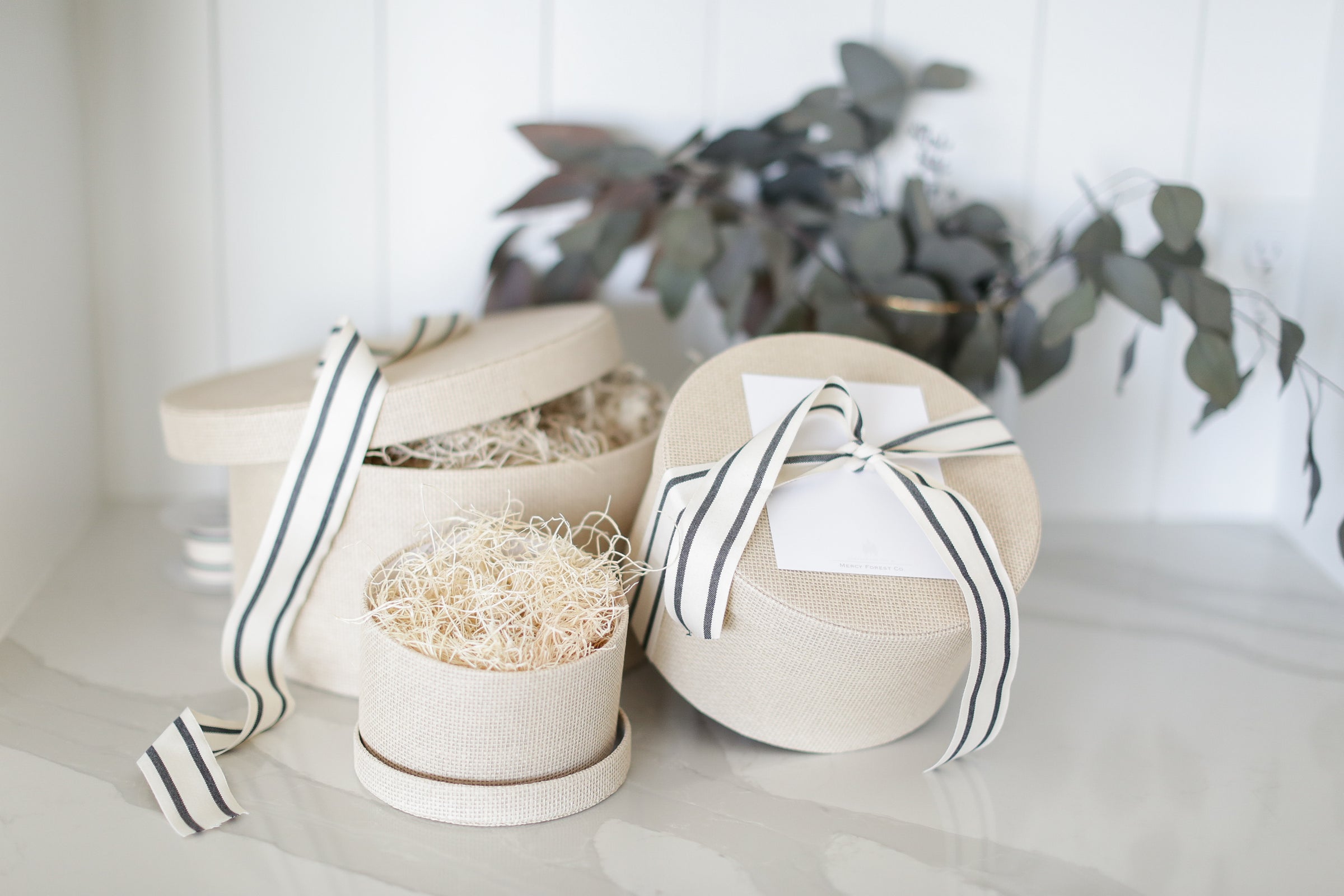Three decorative boxes with ribbons on a light surface with greenery in the background