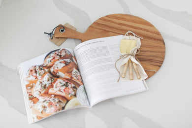 Wood charcuterie board with personalization available, Appetizer and charcuterie cookbook, idea book for charcuterie board, brass and rattan wrapped cheese utensils, white countertop, Mercy Forest Co., custom gifting for realtors, on-demand ready-to-ship gifts, packaged gift box