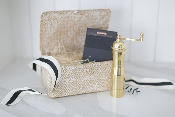 A gold coffee grinder in front of a decorative box with white and black ribbon.
