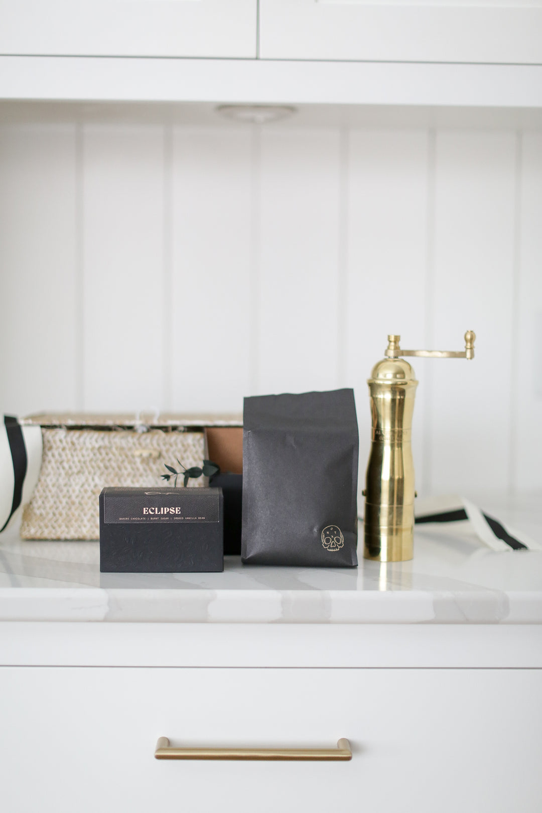 A gold coffee grinder by other coffee items around. Black bags and boxes next to it. Shiplap background.