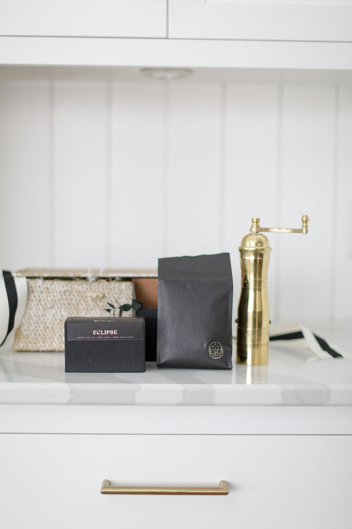 A gold coffee grinder by other coffee items around. Black bags and boxes next to it. Shiplap background.