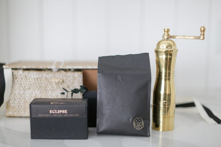 A gold coffee grinder by a black box and bag. other decretive items in the background. shiplap background.
