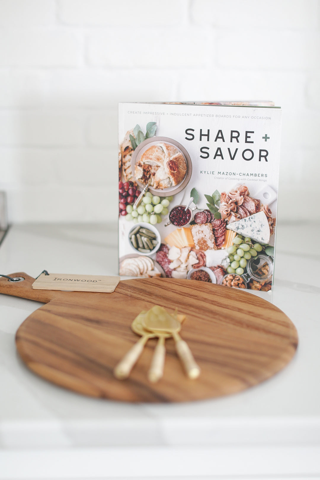 Wood charcuterie board with personalization available, Appetizer and charcuterie cookbook, idea book for charcuterie board, brass and rattan wrapped cheese utensils, white countertop, Mercy Forest Co., custom gifting for realtors, on-demand ready-to-ship gifts, packaged gift box, share and savor cookbook