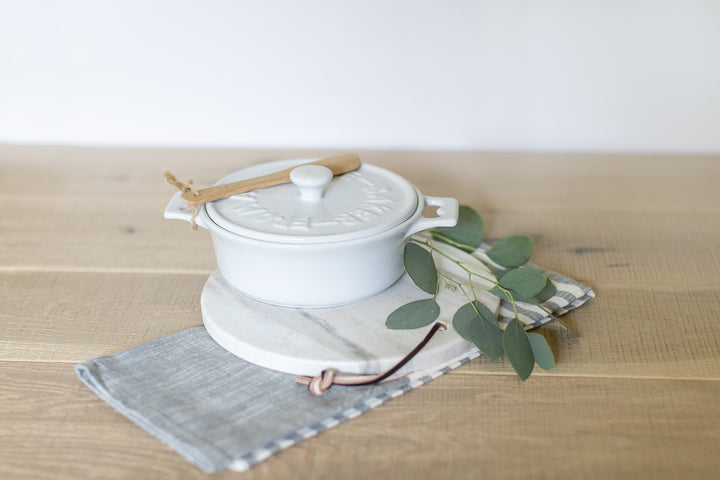White lidded brie baker with wooden spoon on a white and grey marble cutting board and a linen tea towel on a light wood table. Housewarming gifts. Realtor gifts. Mercy Forest Co.