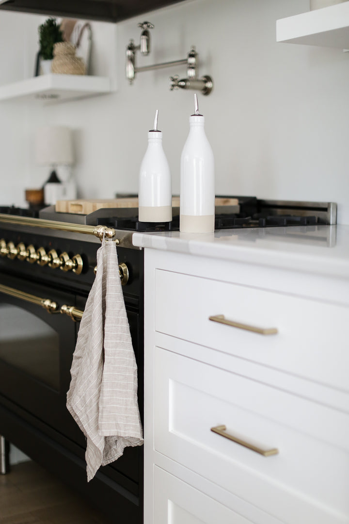 2 white oil cruet's in a kitchen. A grey and white striped tea towel hanging on the oven.